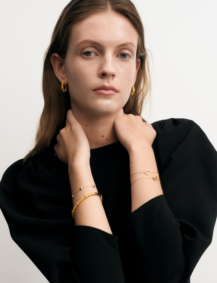 A woman wearing delicate gold bracelets and matching gold hoop earrings, styled with a black long-sleeve top against a plain light gray background.